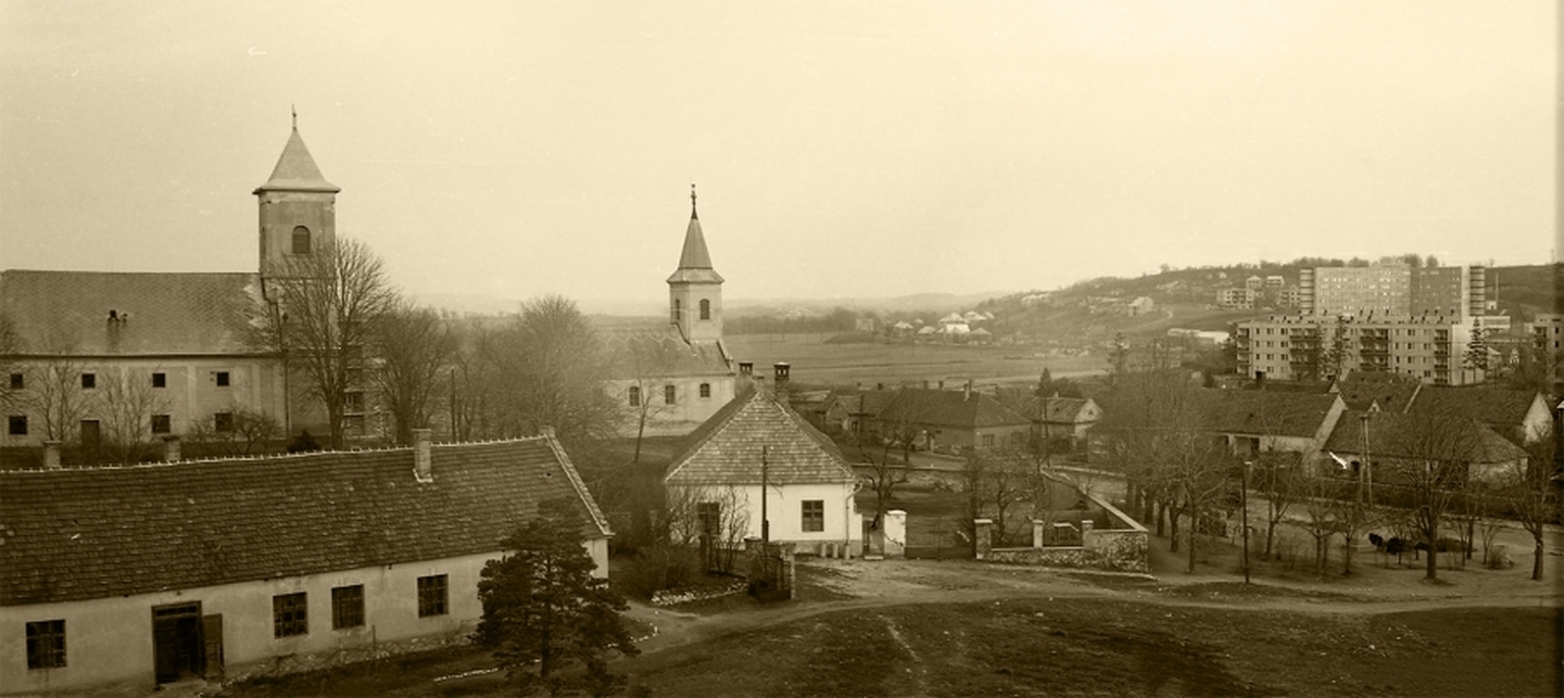 Ajka belvárosának látványa a Templomdombról az 1970-es évek elején. A fotó egyesített, felújított változatán jól látszik a kórház és az első panel épületek sziluettje, valamint a szölőhegy szinte üres oldala. / Ajkai Fotóklub, Sipos Ottó