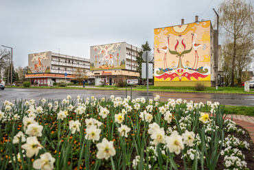 Az 1970-es évek elején épült városközponti tömbben 98 kétszobás lakás mellett földszinti üzletek is helyet kaptak. Főutcai homlokzataikra Darvas Iván helyi festőművész saját kezűleg készített művei 2015 és 2019 között kerültek fel.