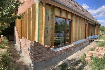 Légtömör épület külső falai, légzáró burok kivitelezése és a lábazati szigetelés beépítése után. Látható az épülő külső burkolótégla előtétfala, és az előtétfalat tartó bekötőpálcák és azok légzáró átvezetése a légzáró, párafékező fólián. A befújt cellulóz hőszigetelést elválasztó fa építőlemez sávok.
